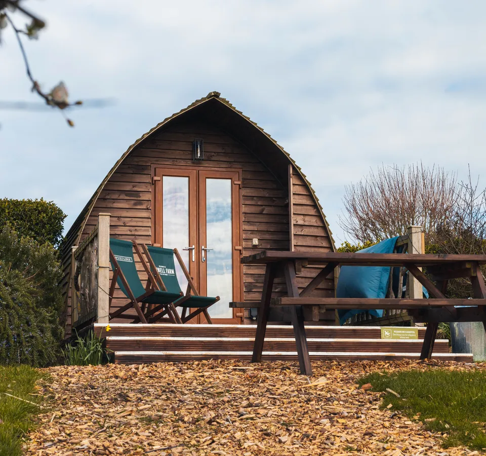 Wooden pod cabin with a small deck, two folding chairs and a picnic table in a garden setting