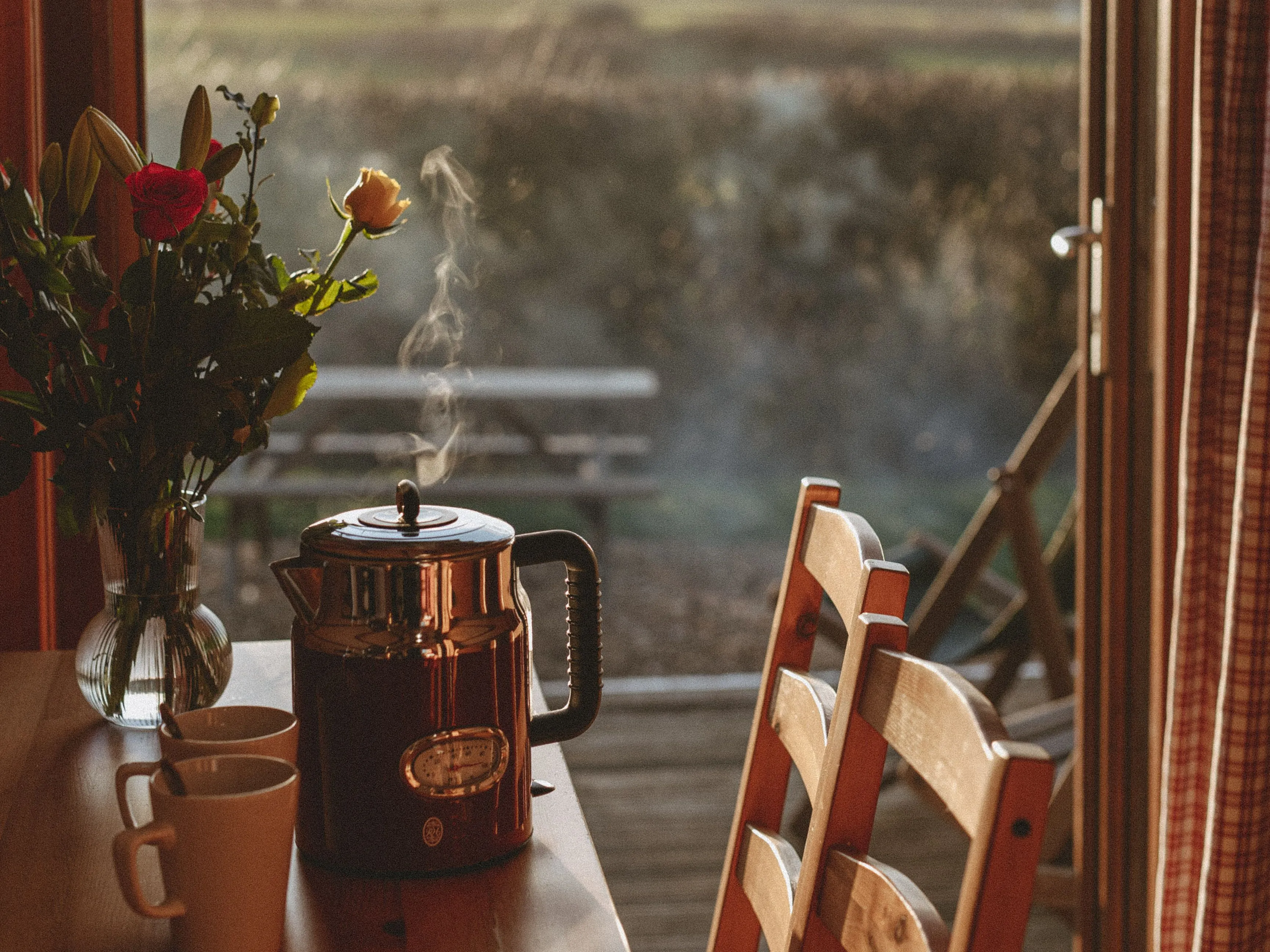 Sunlit table with steaming kettle, mugs and flowers by an open window overlooking countryside