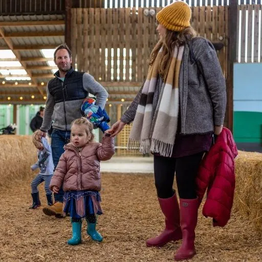 Family walking through a barn play area with hay bales, holding a toddler’s hand