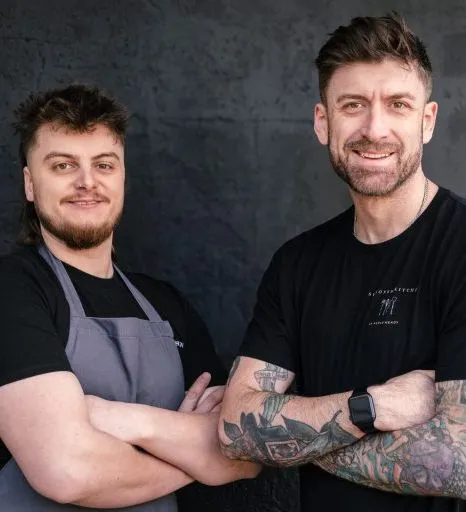Two men standing with arms crossed against a dark wall, one wearing a grey apron and the other showing tattooed arms.