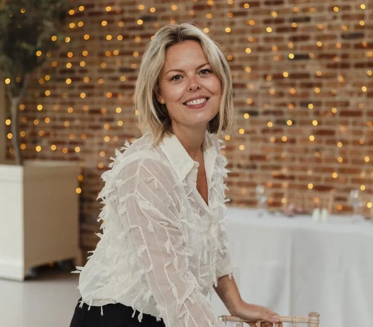 Woman smiling while setting a table at an indoor event, with fairy lights on a brick wall behind her