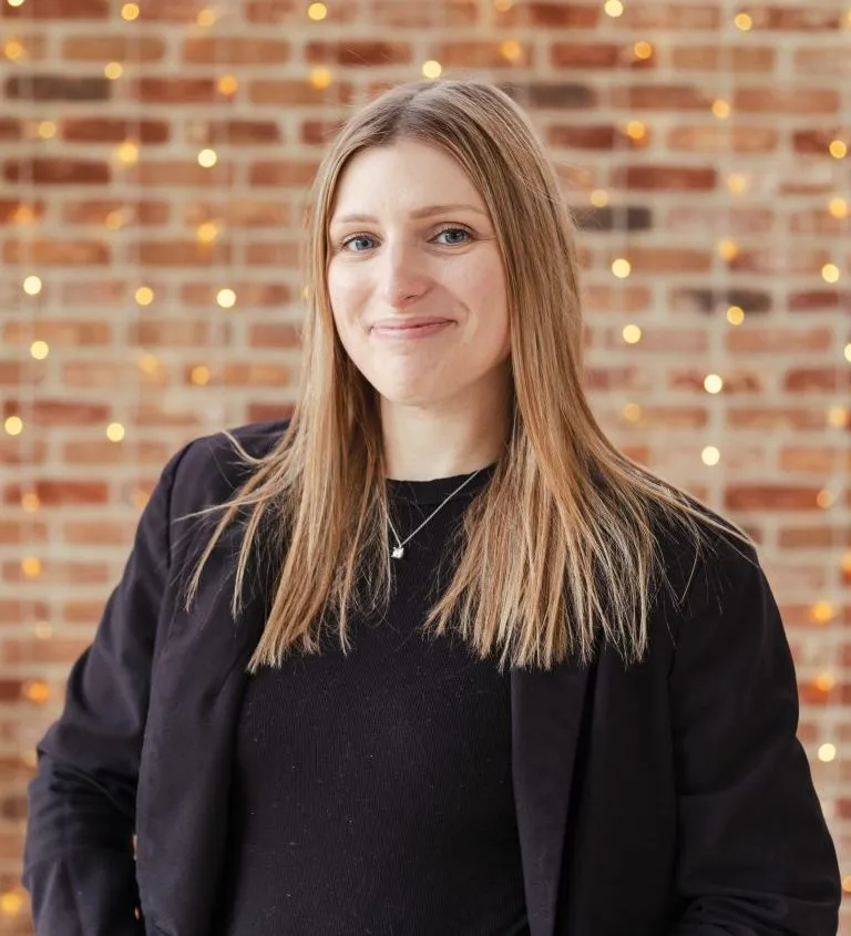 Smiling woman in a black blazer standing in front of a brick wall with warm fairy lights