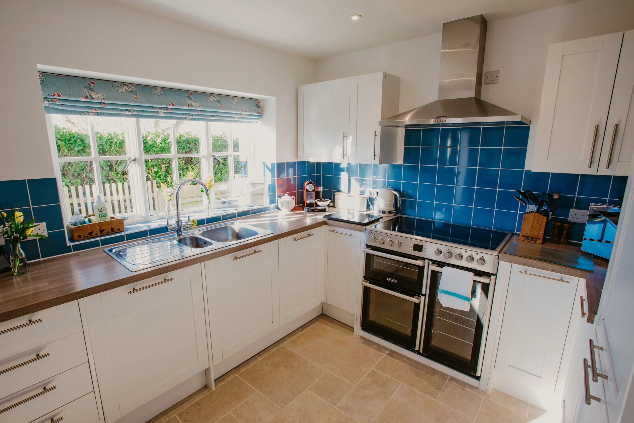 Bright cottage kitchen with white units, blue tiled splashback, range cooker and sink beneath a window