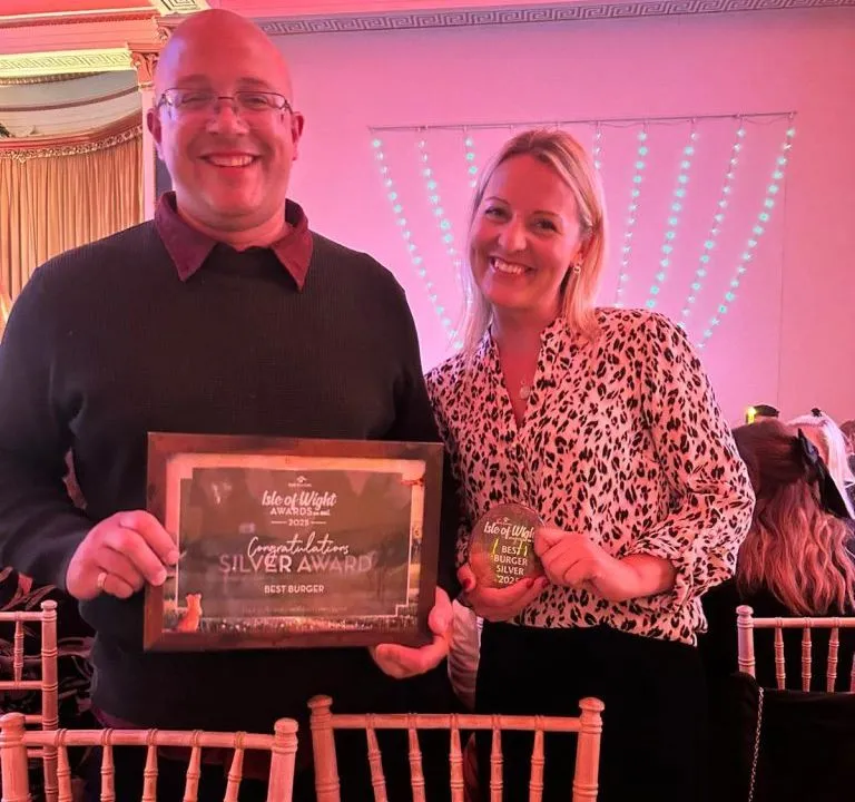 Two people smiling at an awards event, holding a framed Silver Award certificate and a trophy