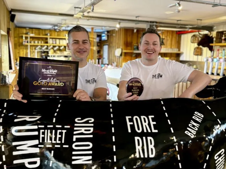 Two men in a restaurant holding a gold award certificate and a burger gold award plaque behind a cow-shaped display