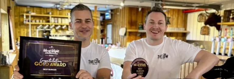 Two men in a restaurant holding a gold award certificate and a burger gold award plaque behind a cow-shaped display