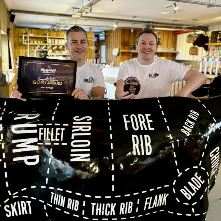 Two men in a restaurant holding a gold award certificate and a burger gold award plaque behind a cow-shaped display