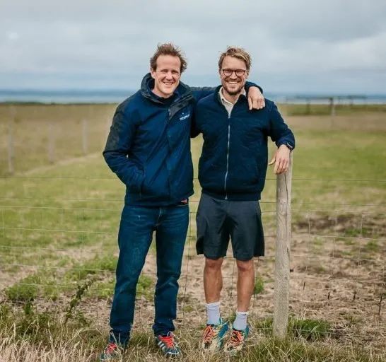 Two men standing arm in arm by a wire fence in a farm field under a cloudy sky.