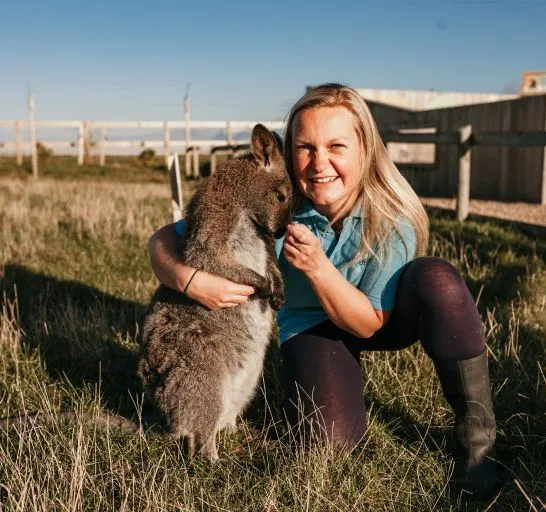 Woman crouching on grass cuddling a wallaby in a paddock at a farm park