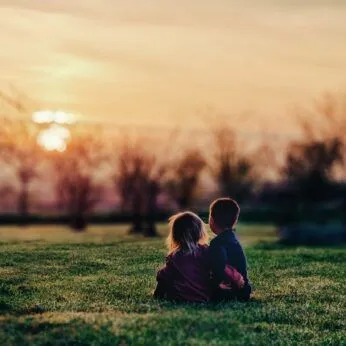Two toddlers sitting on grass watching a sunset near a tent structure