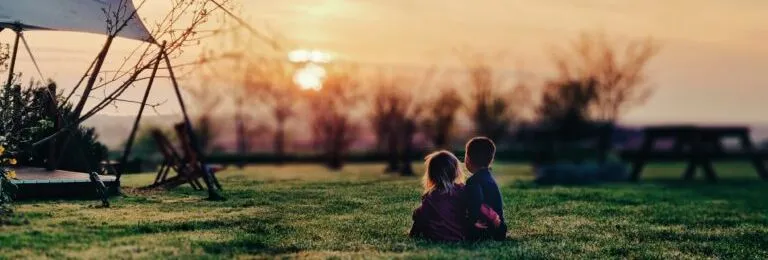 Two toddlers sitting on grass watching a sunset near a tent structure