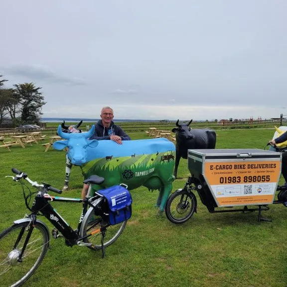 Two men with bicycles beside painted cow sculptures on a grassy field, including an e-cargo bike delivery bicycle