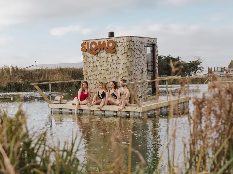 People relaxing on a wooden deck beside a floating Slomo sauna on a lake at Tapnell Farm