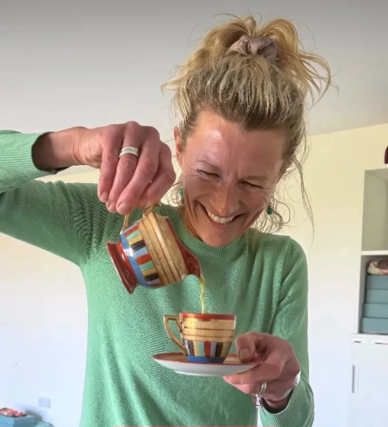 Smiling woman pouring tea from a striped teapot into a matching cup and saucer indoors