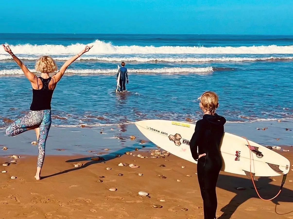 Person practising yoga tree pose on a sandy beach while two surfers in wetsuits head towards the waves with surfboards.