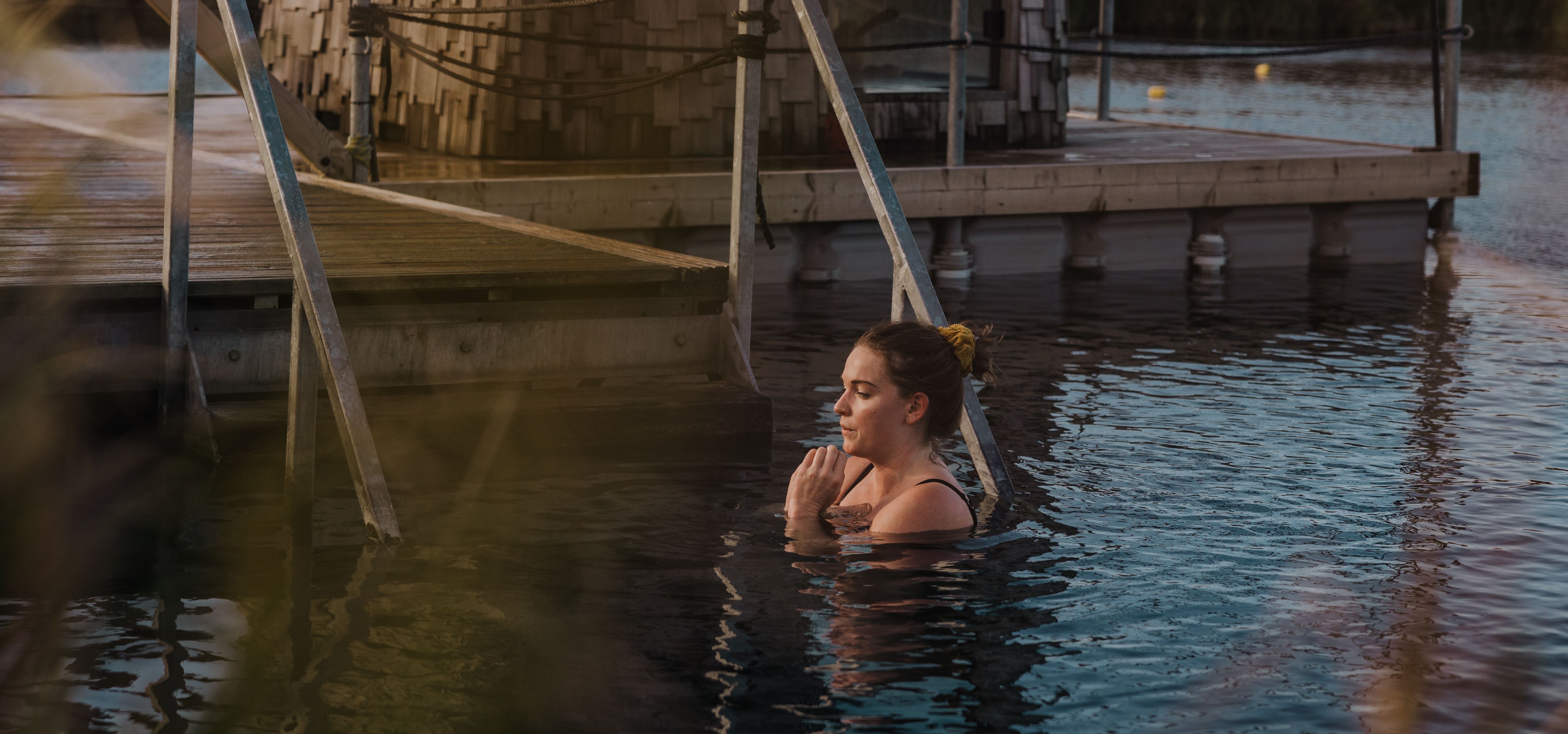 Woman relaxing in a lakeside pool beside a wooden walkway and a shingled building at sunset