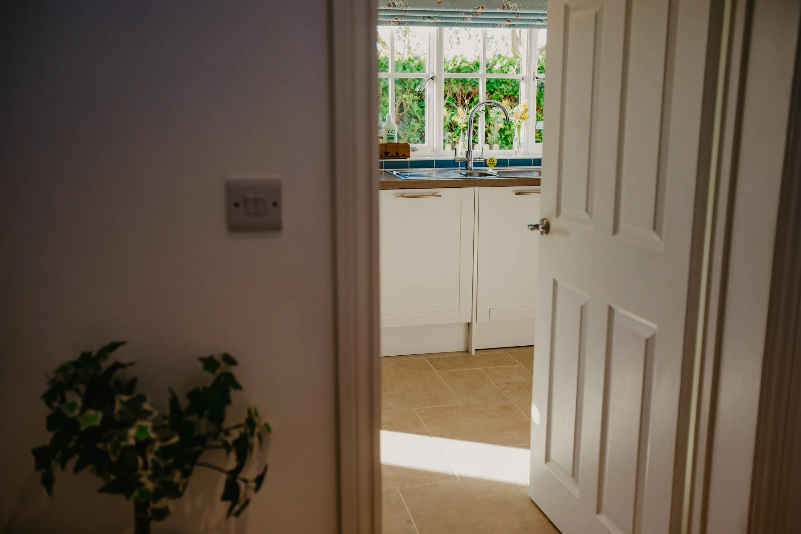 Open door leading into a bright cottage kitchen with sink beneath a window overlooking greenery