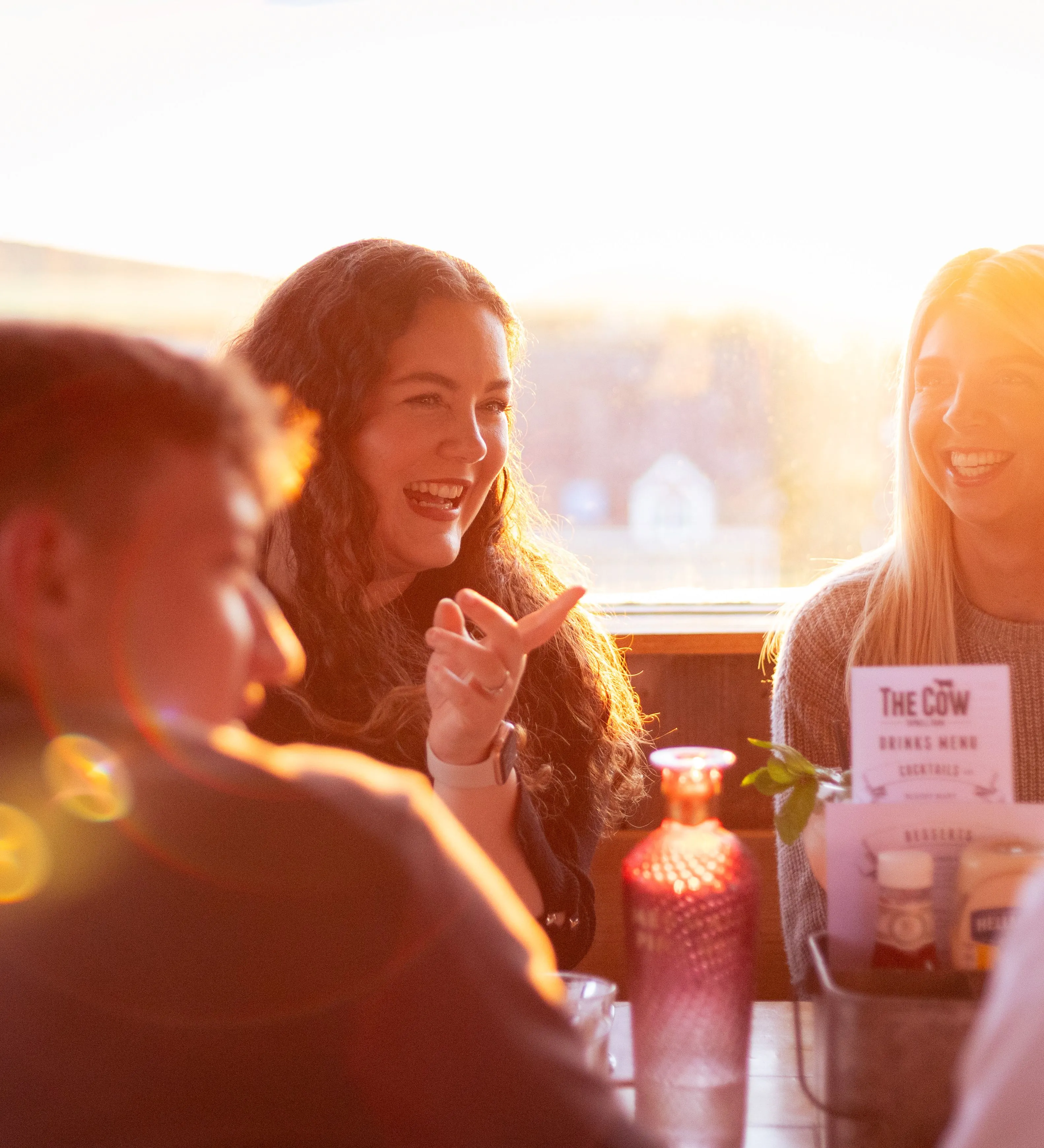 Friends laughing around a table in The Cow restaurant at Tapnell Farm, lit by warm evening sunlight