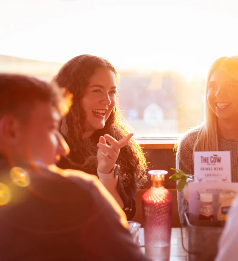 Friends laughing around a table in The Cow restaurant at Tapnell Farm, lit by warm evening sunlight