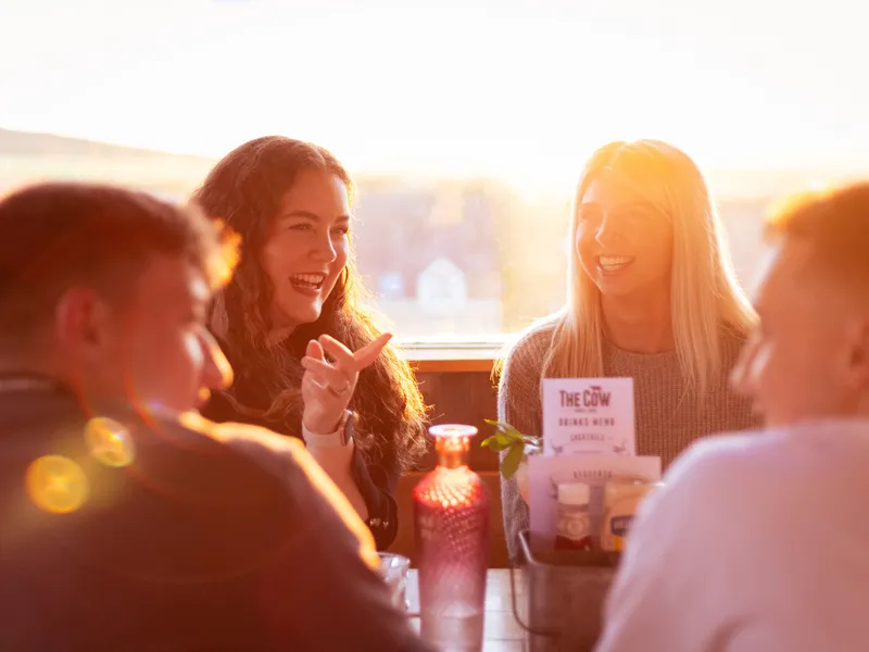 Friends laughing around a table in The Cow restaurant at Tapnell Farm, lit by warm evening sunlight