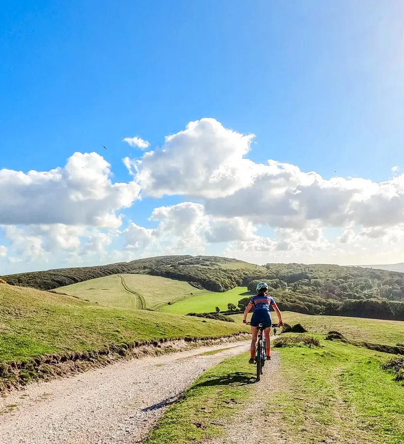 Cyclist riding along a gravel track through rolling green hills under a blue sky with white clouds