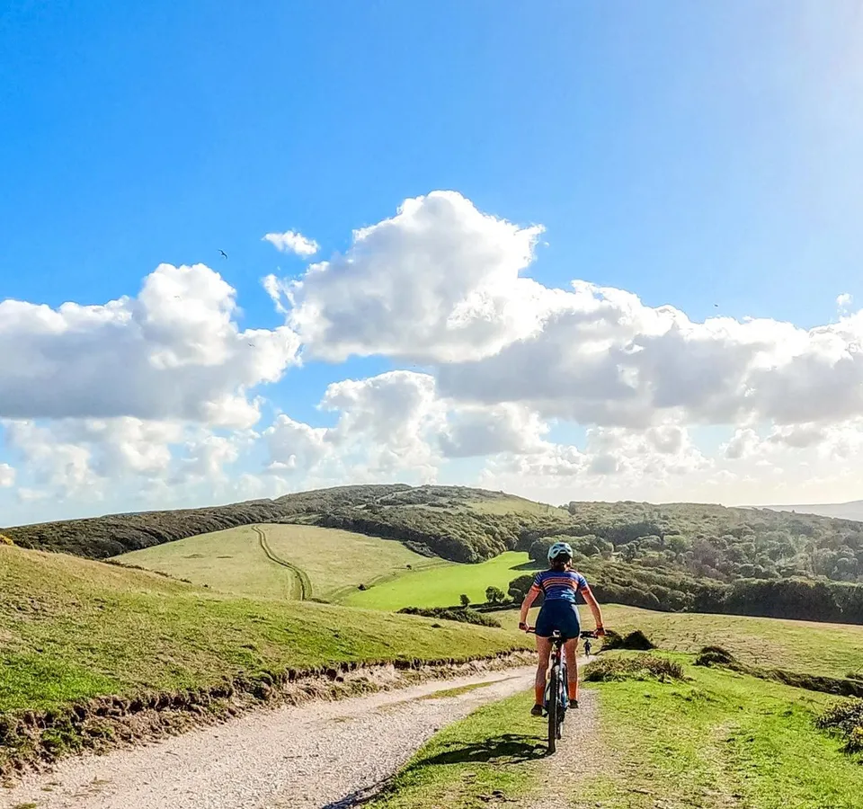 Cyclist riding along a gravel track through rolling green hills under a blue sky with white clouds