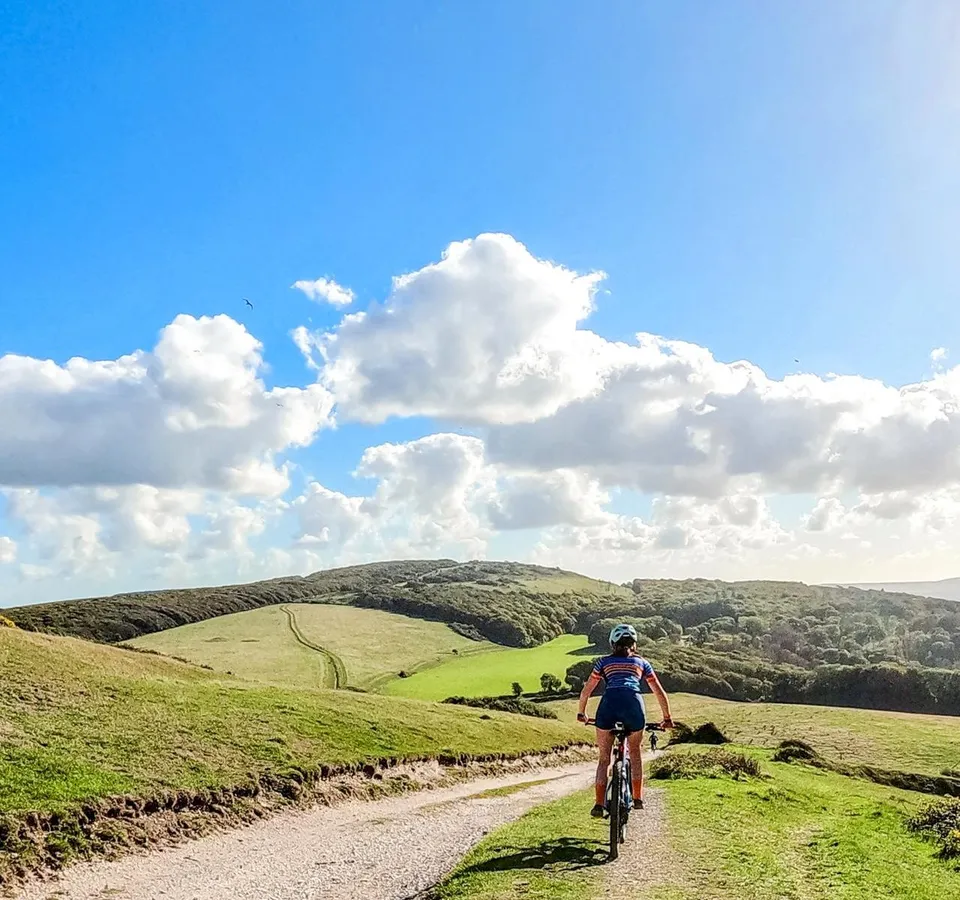 Cyclist riding along a gravel track through rolling green hills under a blue sky with white clouds