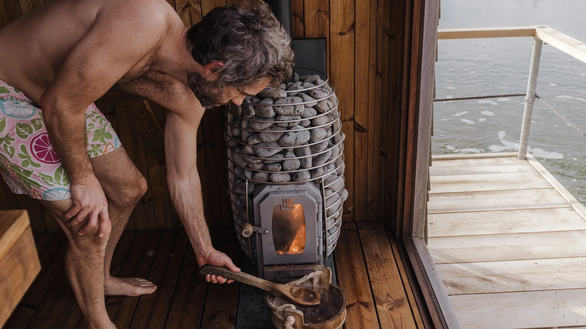 Man tending a wood-fired sauna stove inside a floating sauna, with water visible through the open doorway