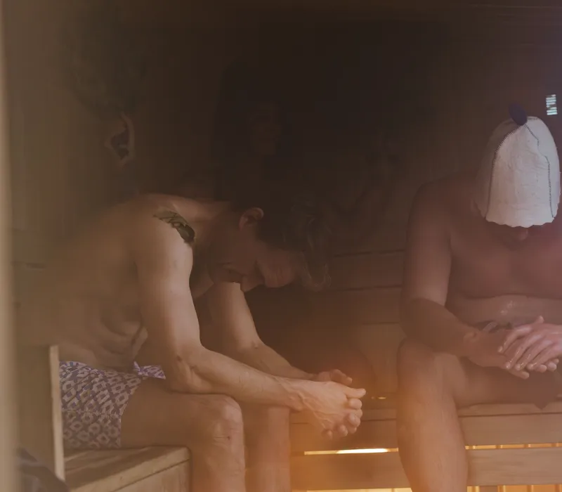Two men sitting in a steamy wooden sauna, one wearing a sauna hat.