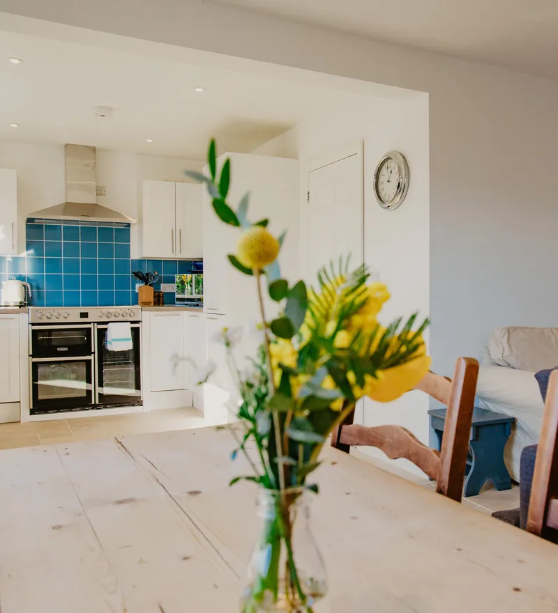 Open-plan cottage kitchen and sitting area with blue tiled splashback, dining table and vase of flowers