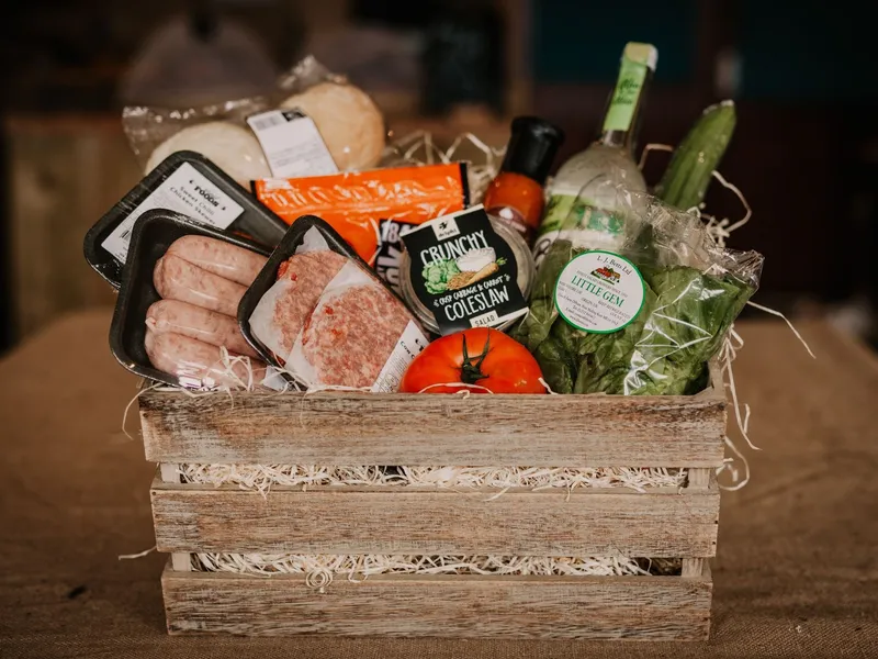 Wooden crate hamper filled with sausages, burgers, rolls, salad leaves, tomato, cucumber, coleslaw and bottles of sauce
