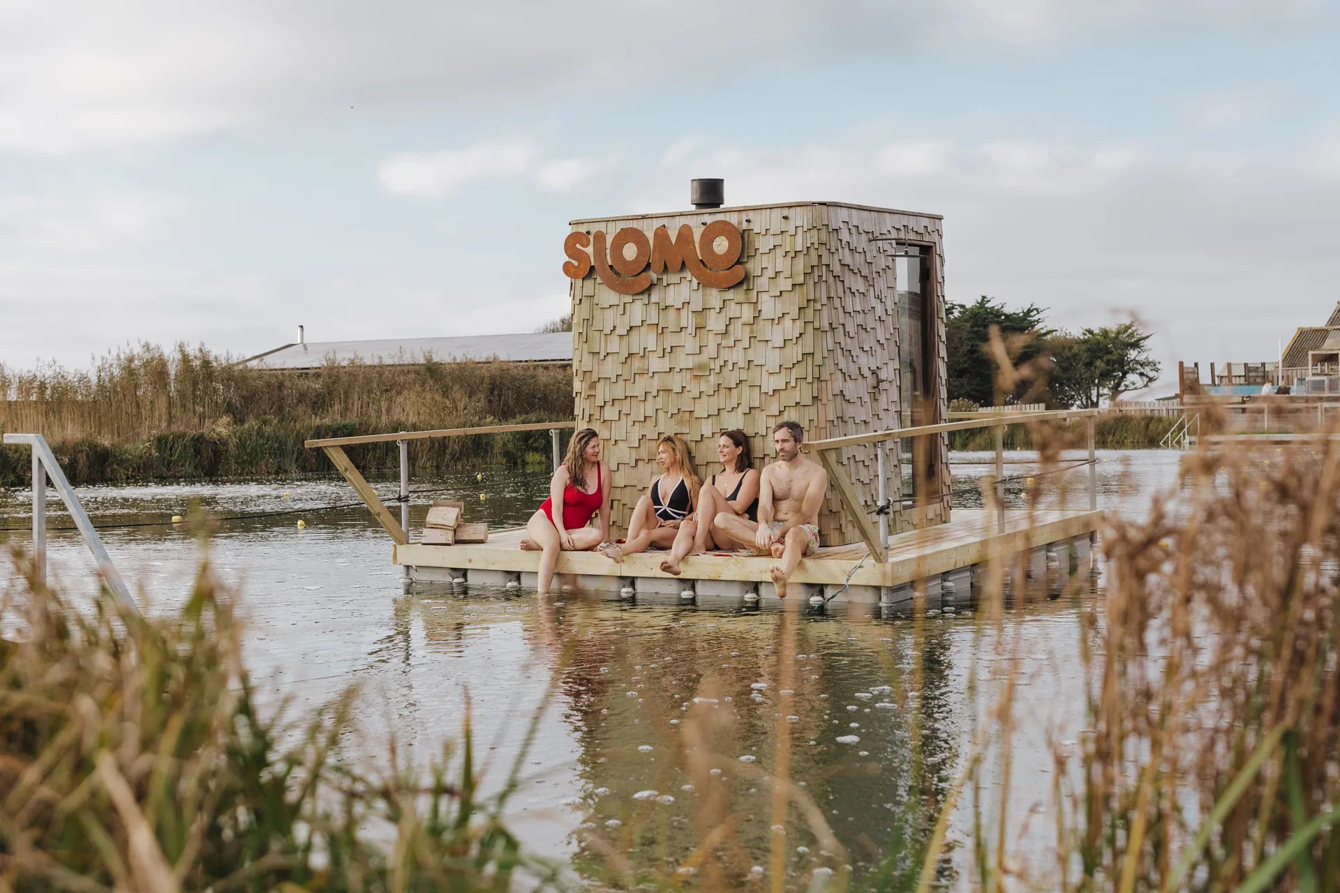 Four people sitting on the deck of a floating sauna hut labeled 'SLOMO' on a tranquil body of water.