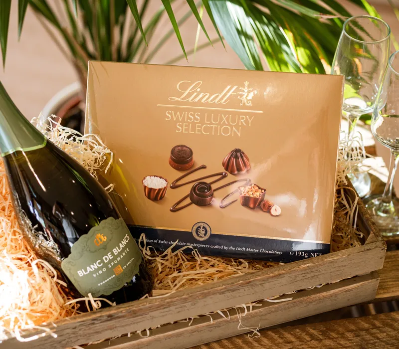 Gift hamper with a bottle of sparkling wine, a Lindt Swiss Luxury Selection chocolate box, and two champagne flutes in a wooden crate