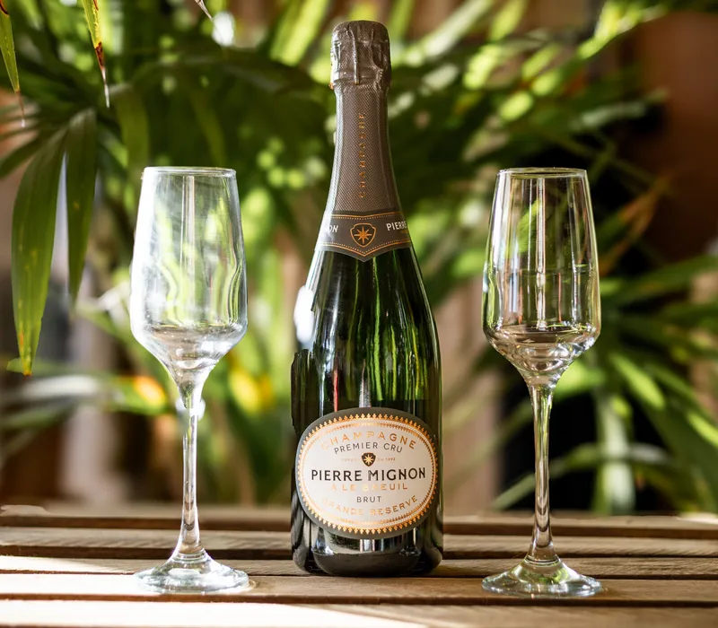 Bottle of Pierre Mignon champagne between two empty flutes on a wooden table with green plants in the background