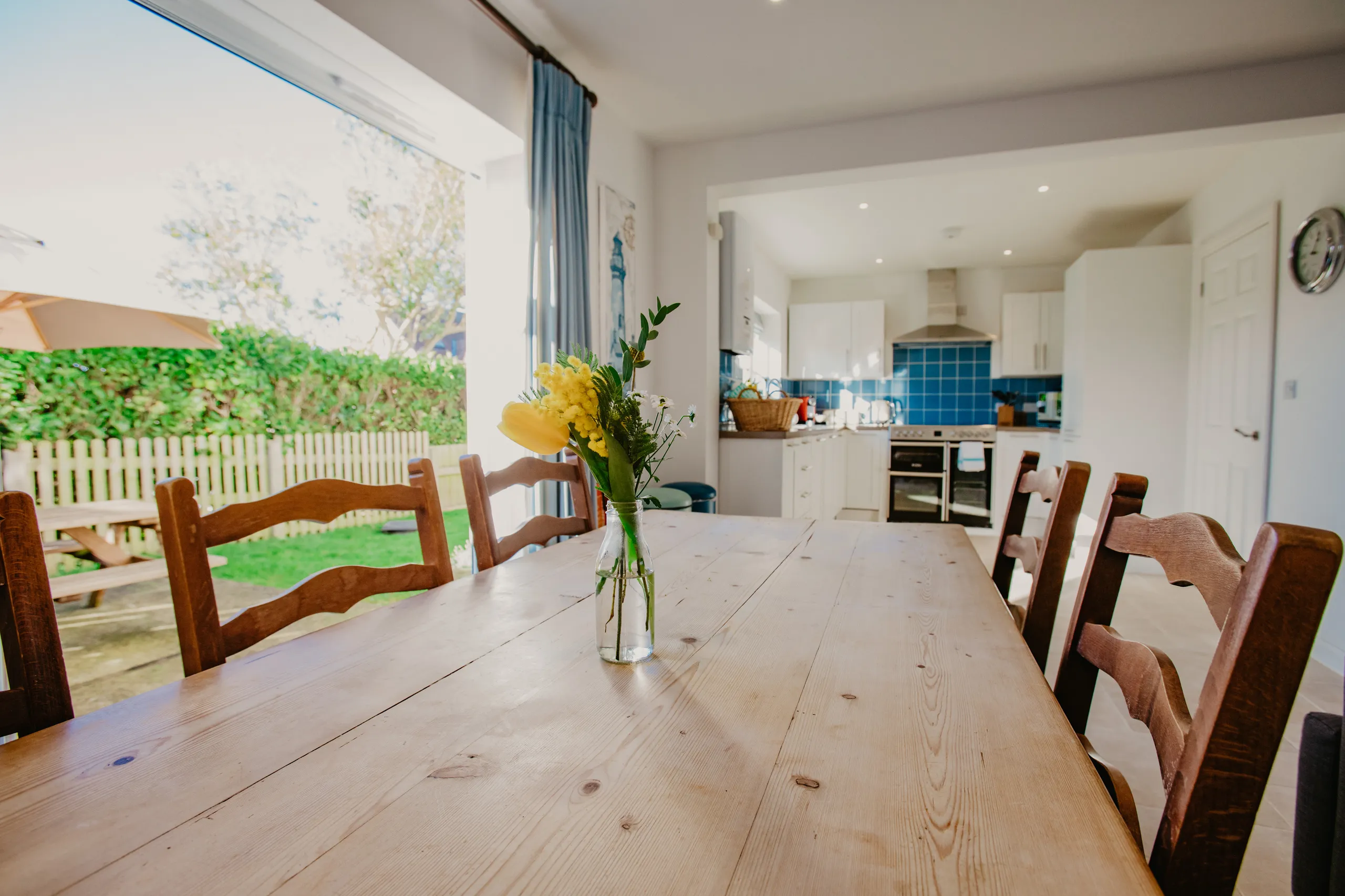 Bright dining area with wooden table and chairs, flowers in a vase, and open-plan kitchen beyond with blue tiled splashback
