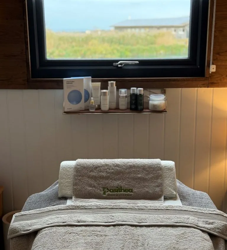 Massage bed with folded towels beneath a window, skincare products on a shelf and countryside view outside