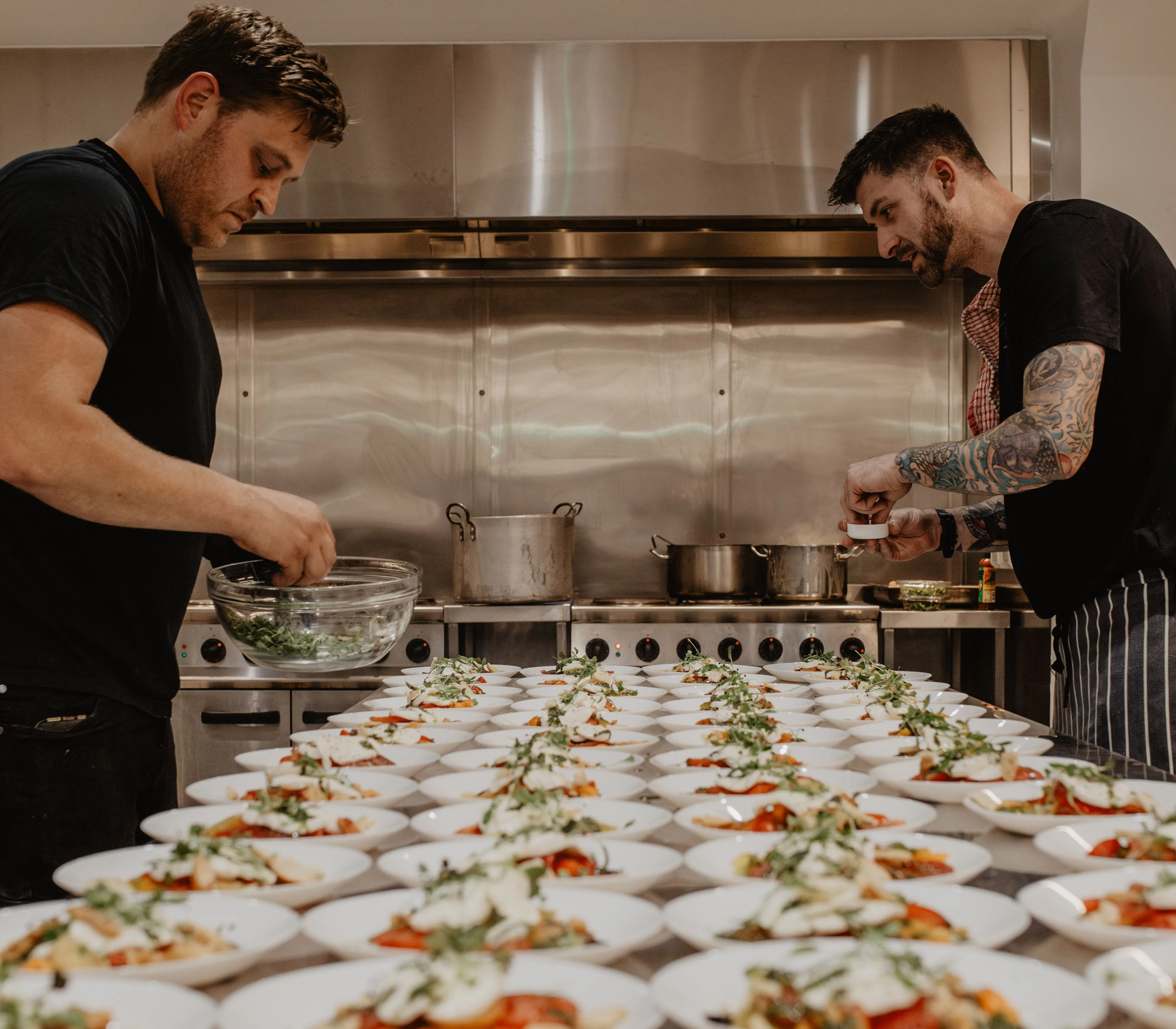 Two chefs plating starters in a professional kitchen with rows of dishes on a long table