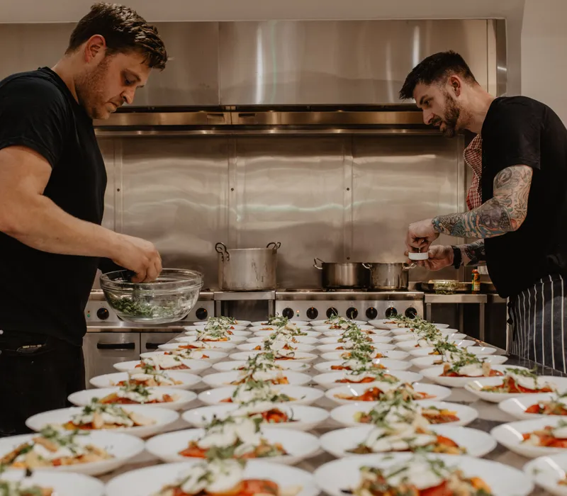 Two chefs plating starters in a professional kitchen with rows of dishes on a long table