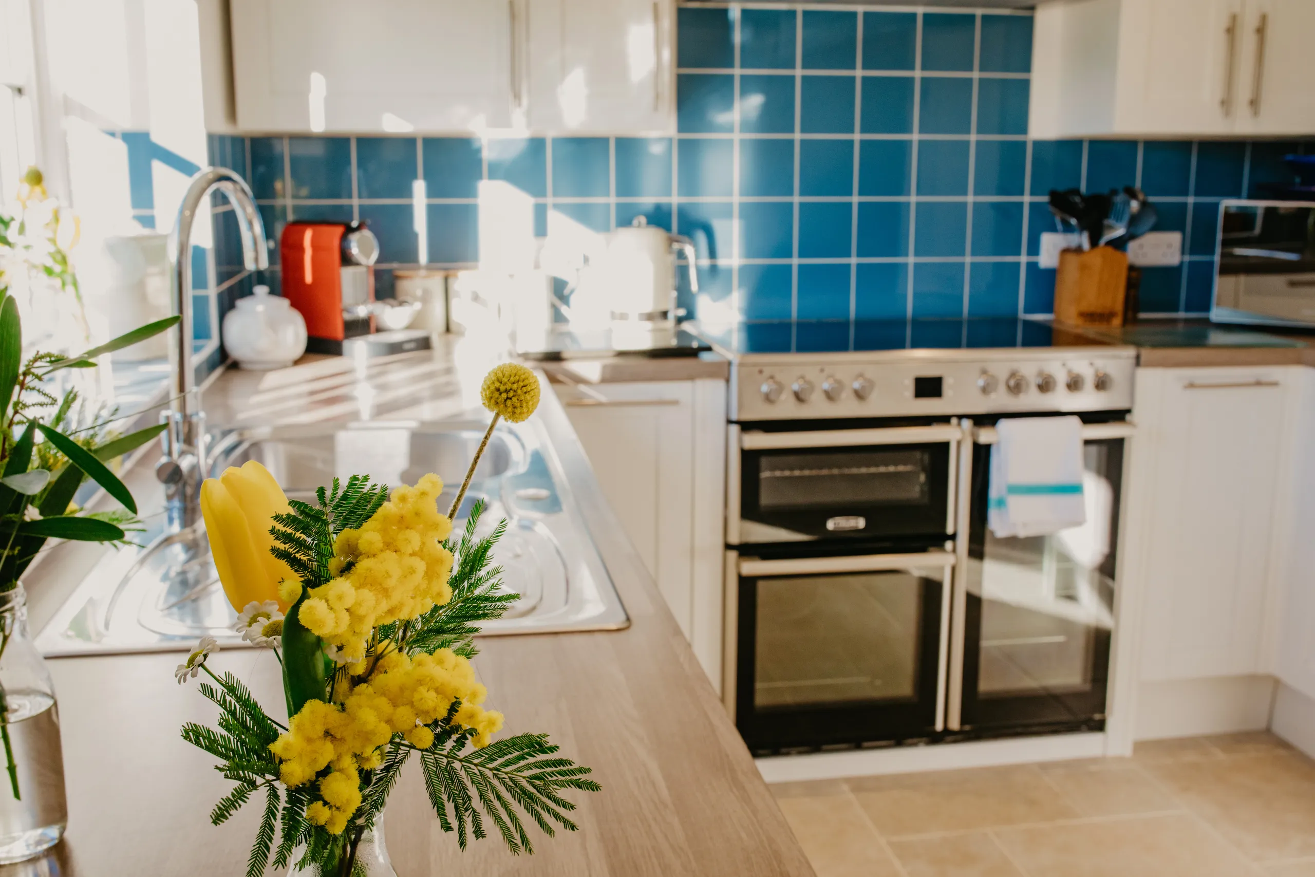 Bright cottage kitchen with blue tiled splashback, white cupboards, double oven and yellow flowers on the worktop