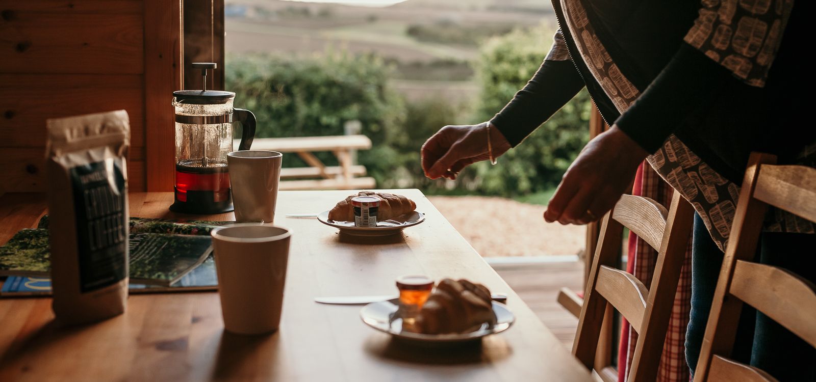 Person arranging breakfast on a wooden table with croissants, mugs and a French press in a cabin with countryside views