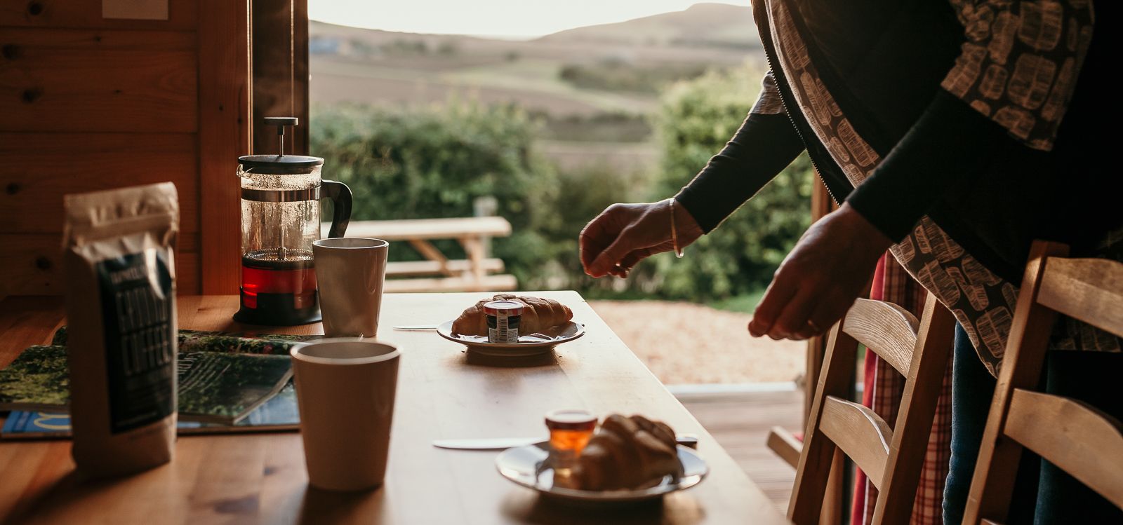 Person arranging breakfast on a wooden table with croissants, mugs and a French press in a cabin with countryside views