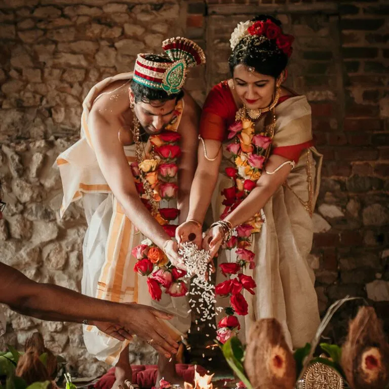 Bride and groom in traditional Indian wedding attire performing a ritual by offering rice into a sacred fire