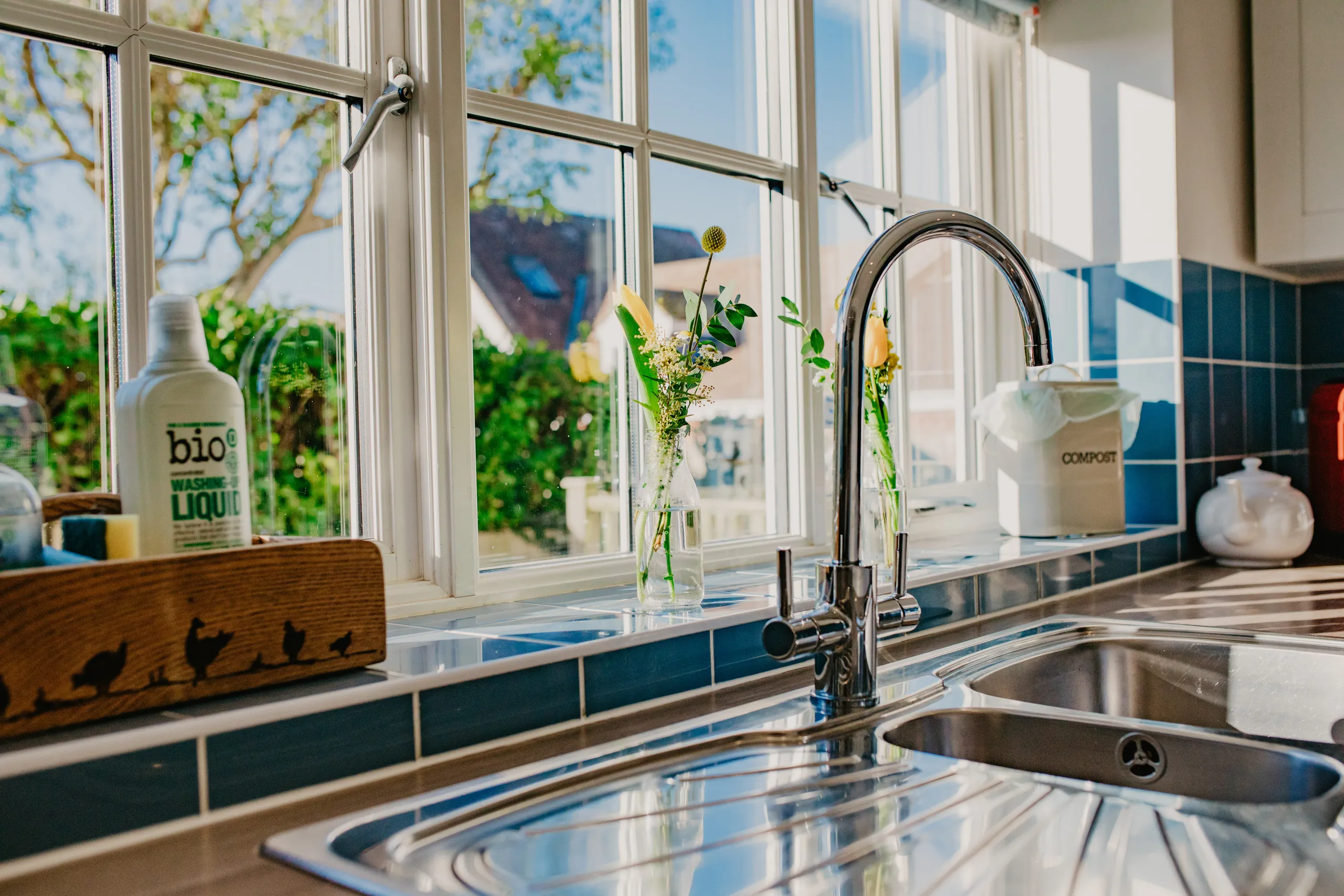 Sunlit kitchen sink with chrome tap, blue tiled splashback and flowers on the windowsill