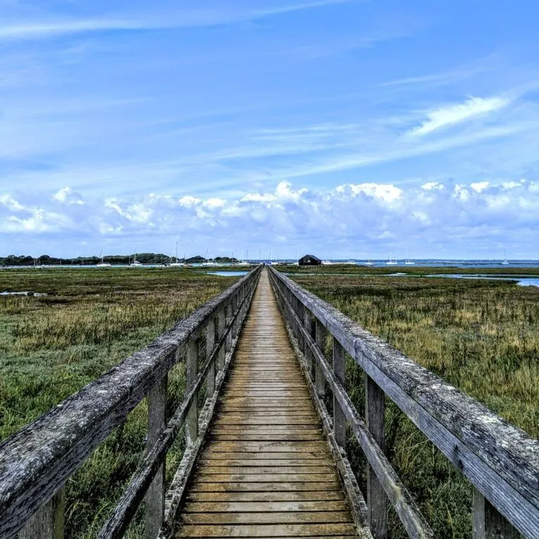 Wooden boardwalk stretching across salt marsh towards distant buildings under a blue, cloud-streaked sky