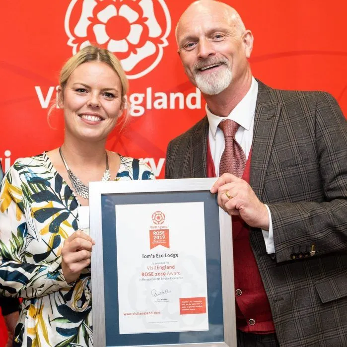 Two people holding a framed VisitEngland ROSE 2019 award certificate in front of a red VisitEngland backdrop