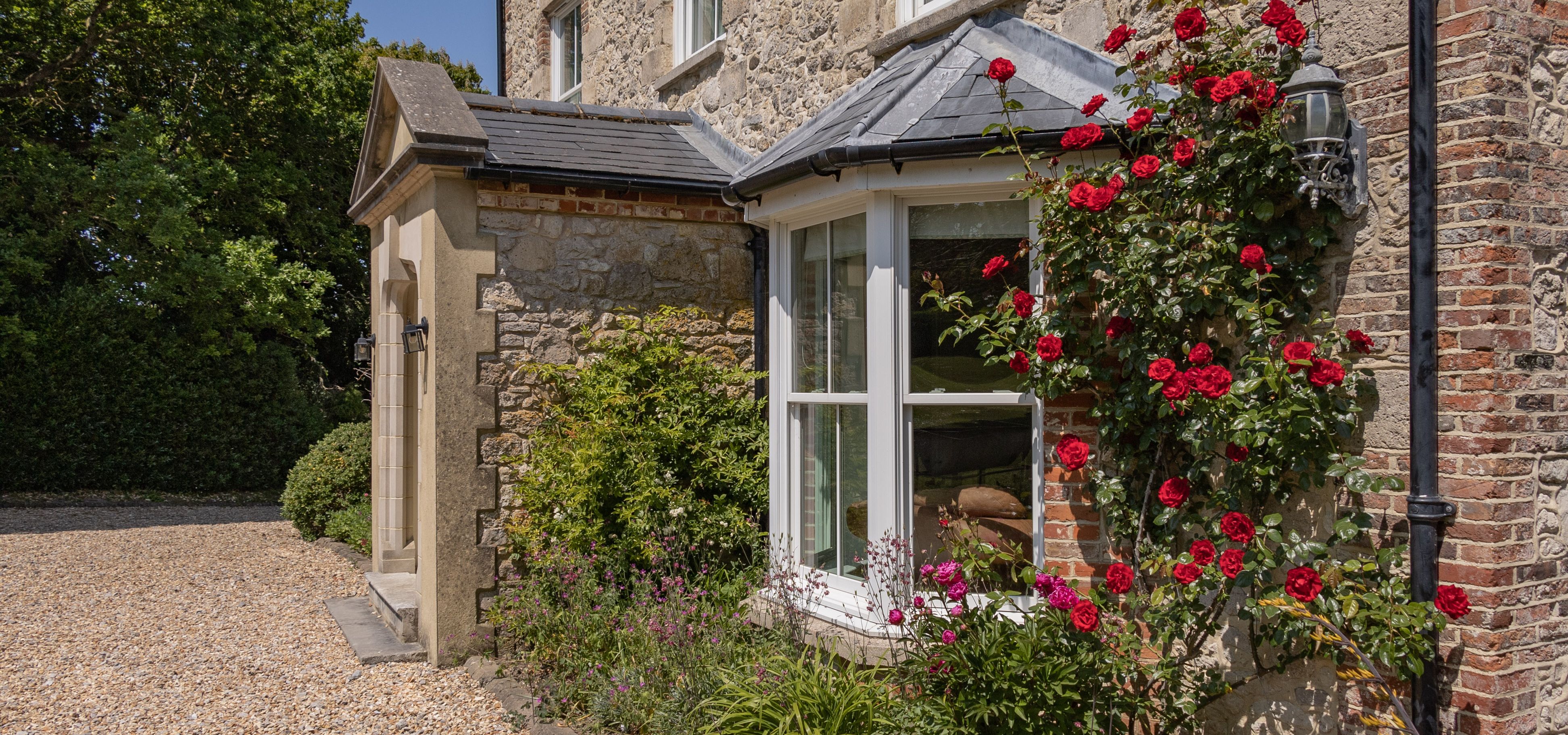 Stone farmhouse exterior with bay window and red climbing roses beside a gravel path