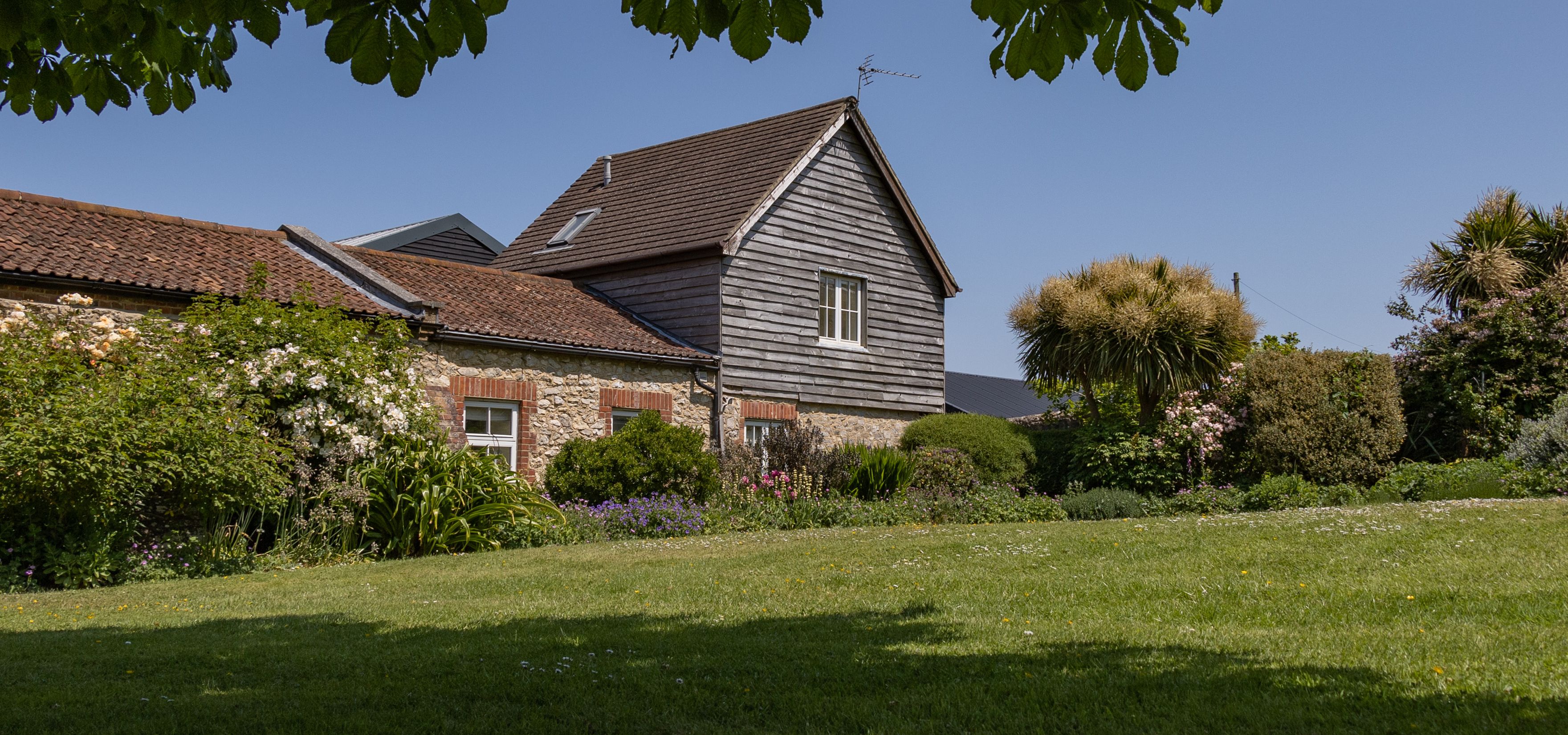East Afton Farmhouse with stone walls and timber-clad gable, seen across a lawn and flower-filled garden under leafy branches