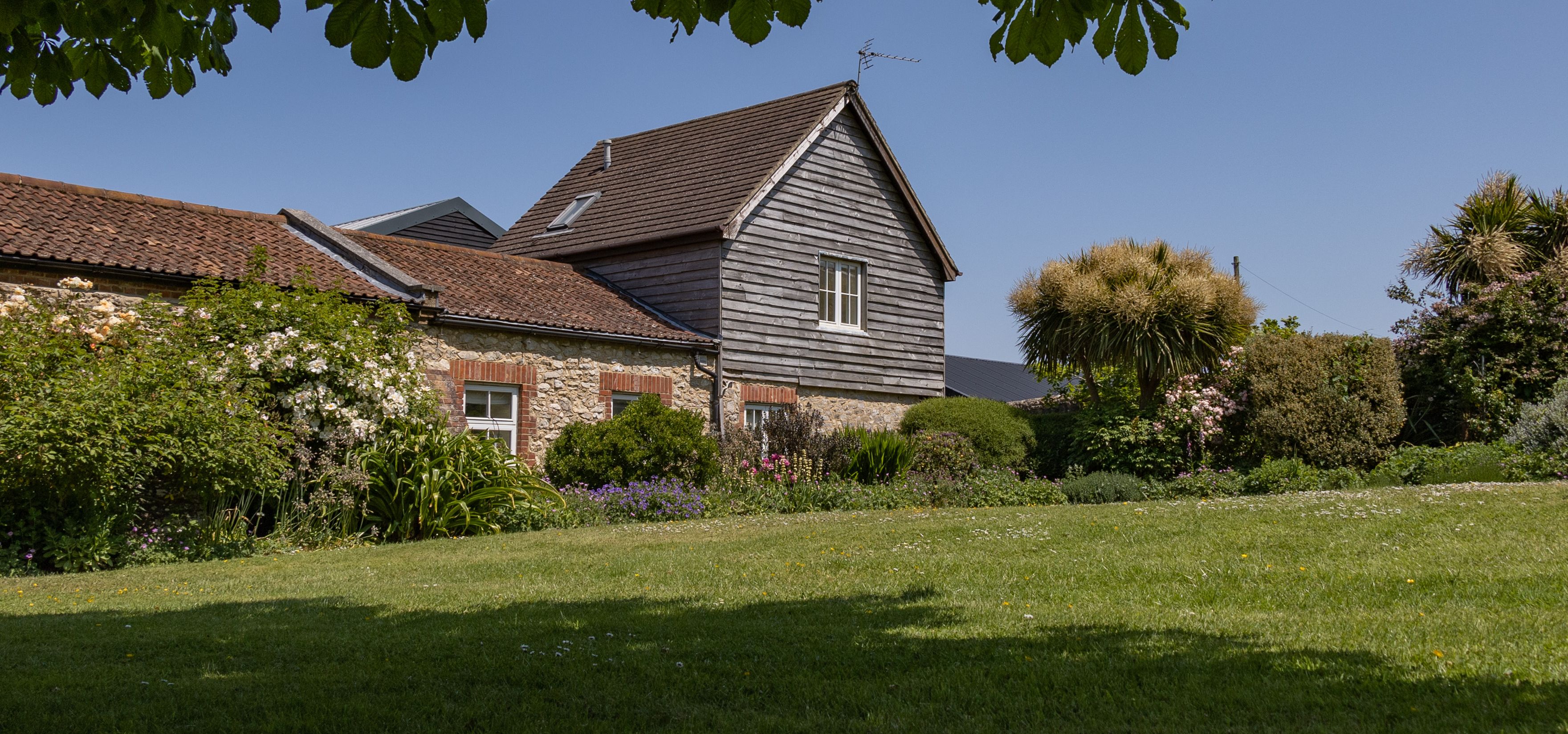 East Afton Farmhouse with stone walls and timber-clad gable, seen across a lawn and flower-filled garden under leafy branches