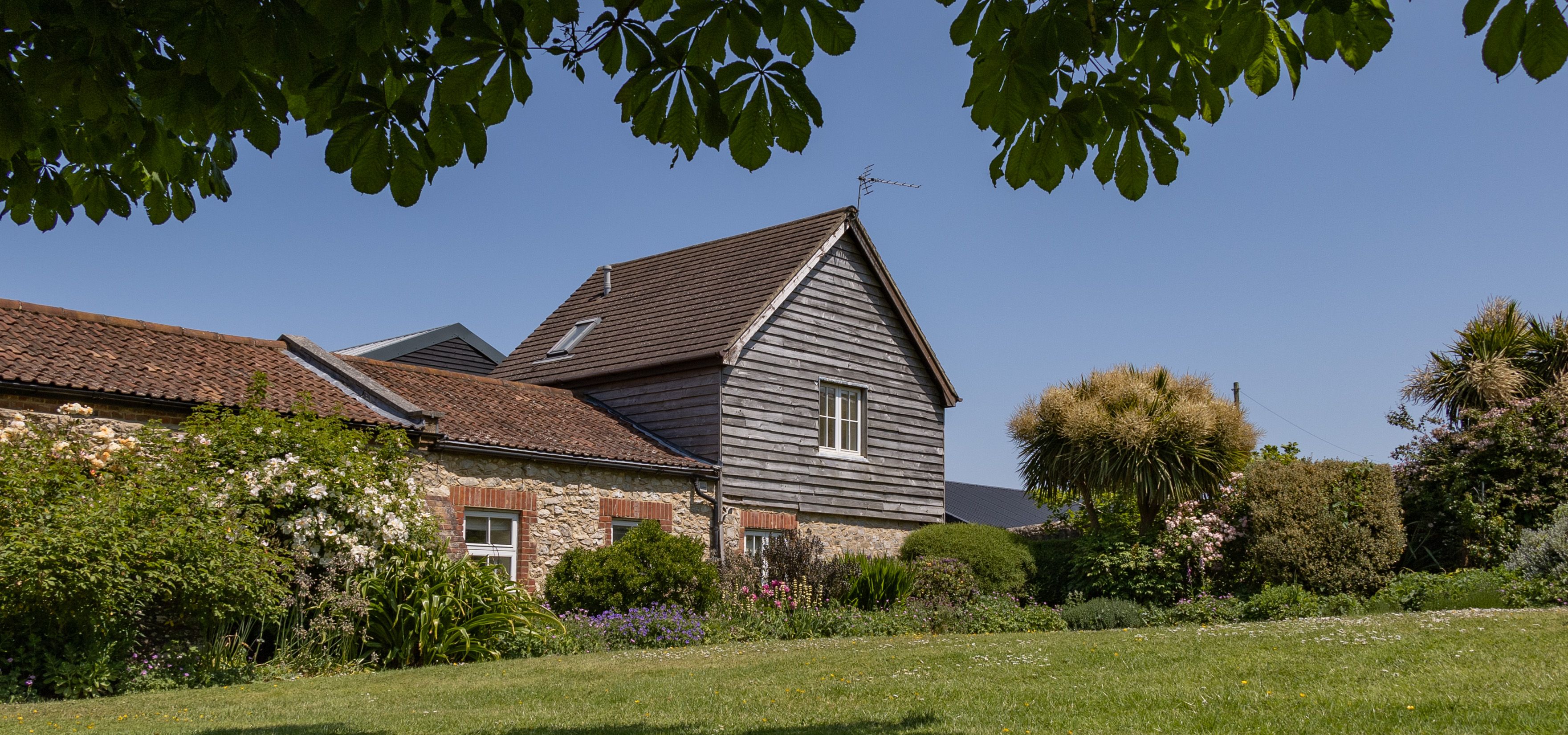 East Afton Farmhouse with stone walls and timber-clad gable, seen across a lawn and flower-filled garden under leafy branches