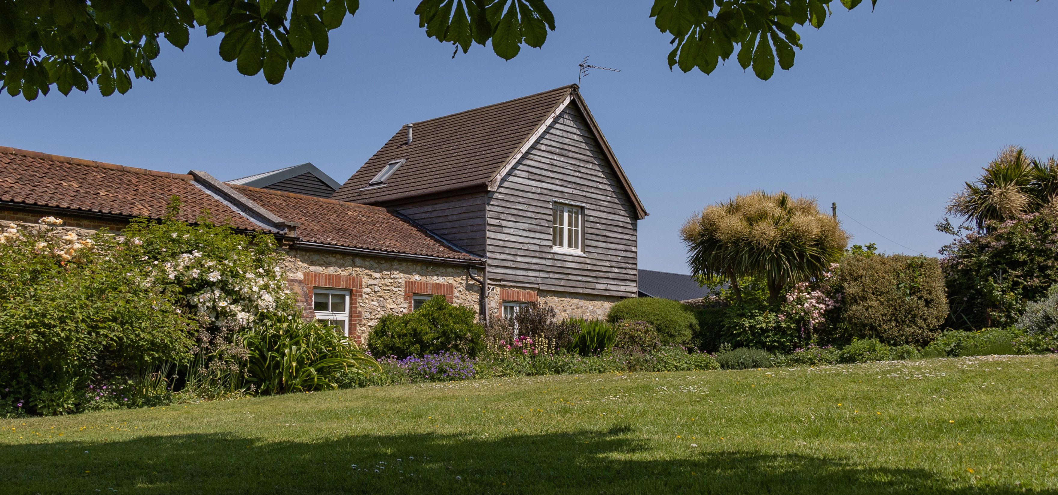 East Afton Farmhouse with stone walls and timber-clad gable, seen across a lawn and flower-filled garden under leafy branches